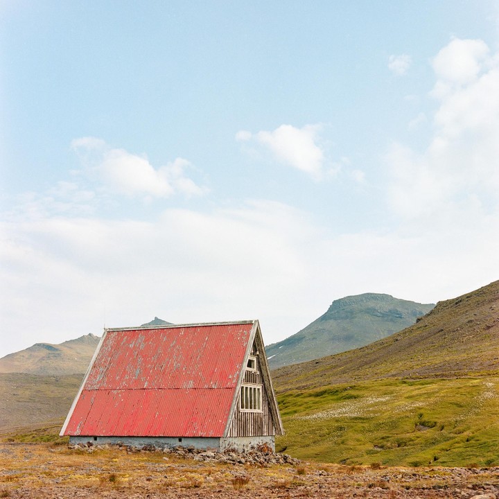 An A-frame cabin with a red roof sits in a vast, rocky landscape with mountains in the background.