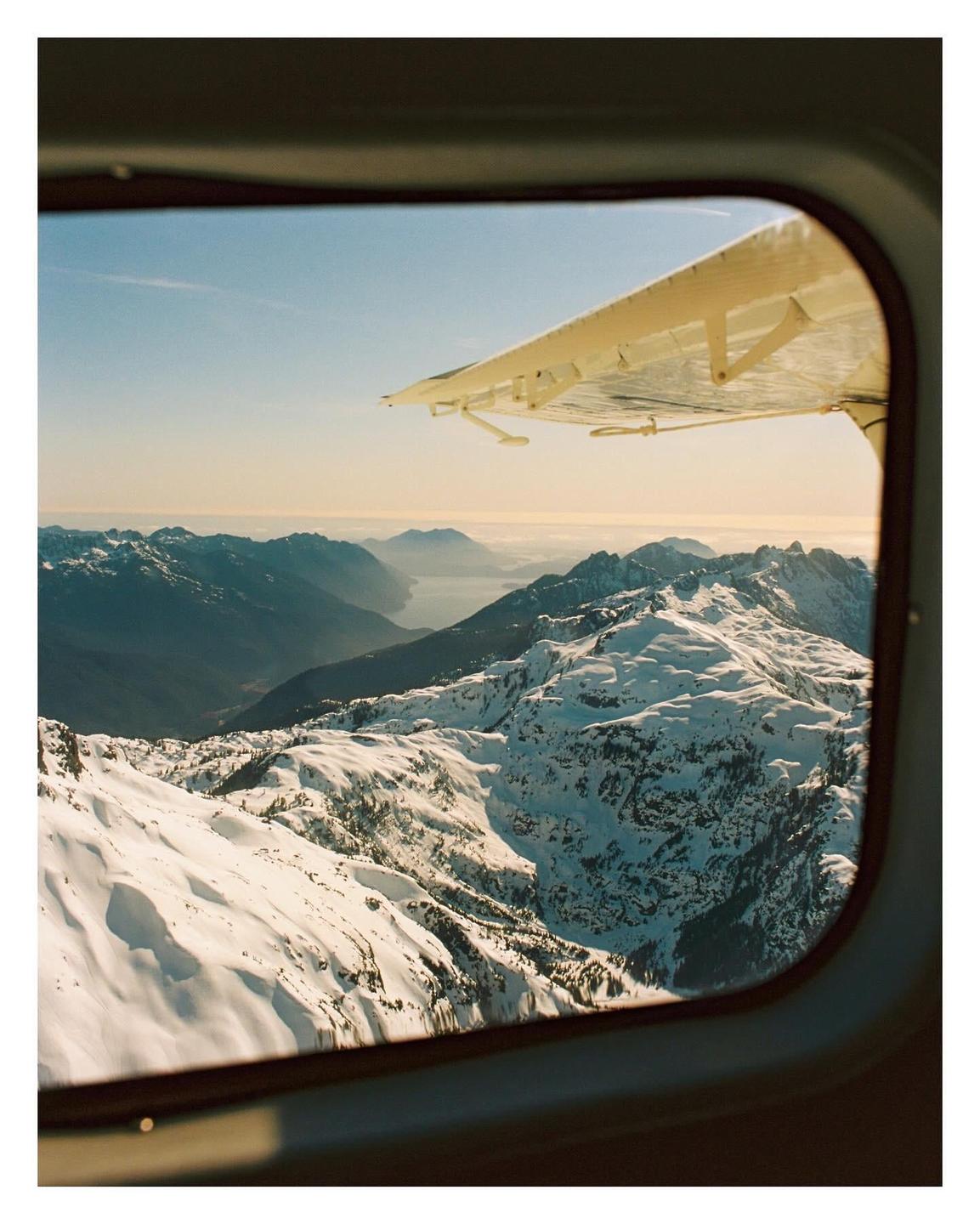 A view from an airplane window shows snow-covered mountains and a body of water in the distance.