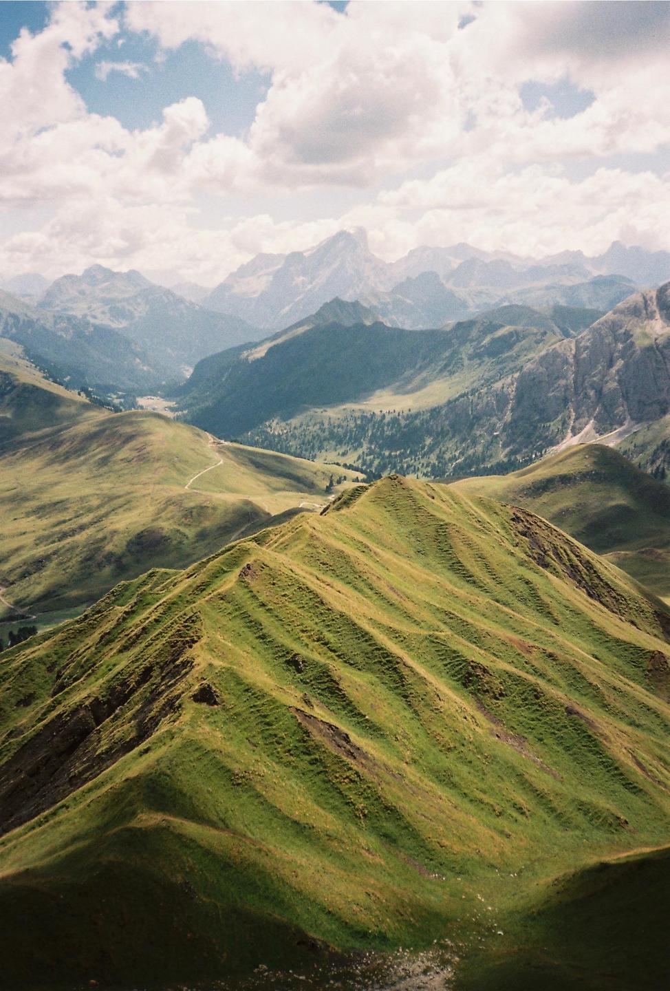 A winding path leads through a valley surrounded by green, rolling hills and distant mountains under a cloudy sky.