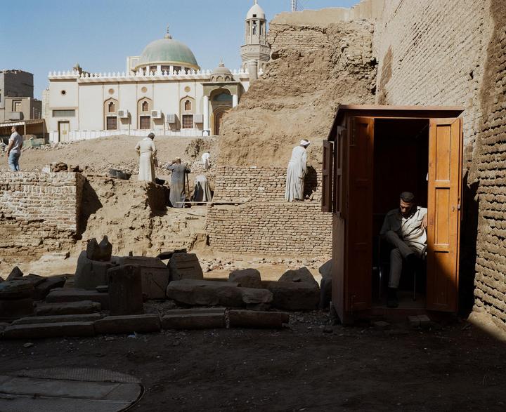 A man sits in a wooden booth while other men work at an excavation site with a mosque in the background.
