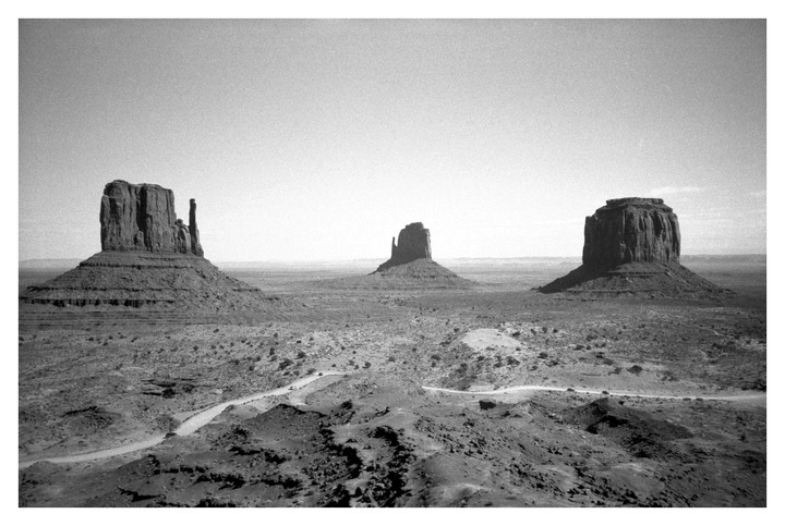 Three large rock formations stand in a vast, arid landscape with a winding dirt road in the foreground.