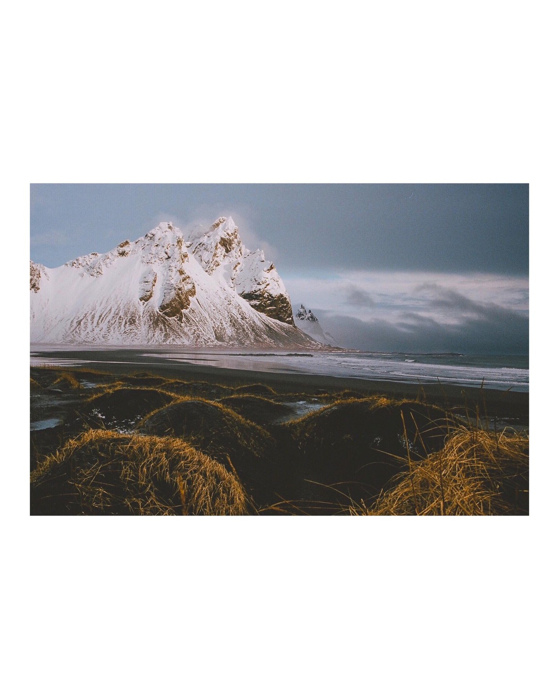 A snow-covered mountain rises above a dark beach with waves crashing on the shore and tufts of dry grass in the foreground.