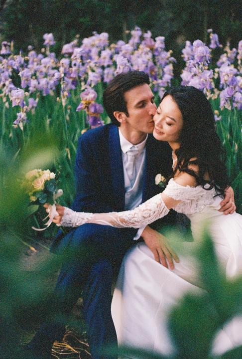 A man in a suit kisses a woman in a wedding dress on the cheek while sitting in a field of purple flowers.