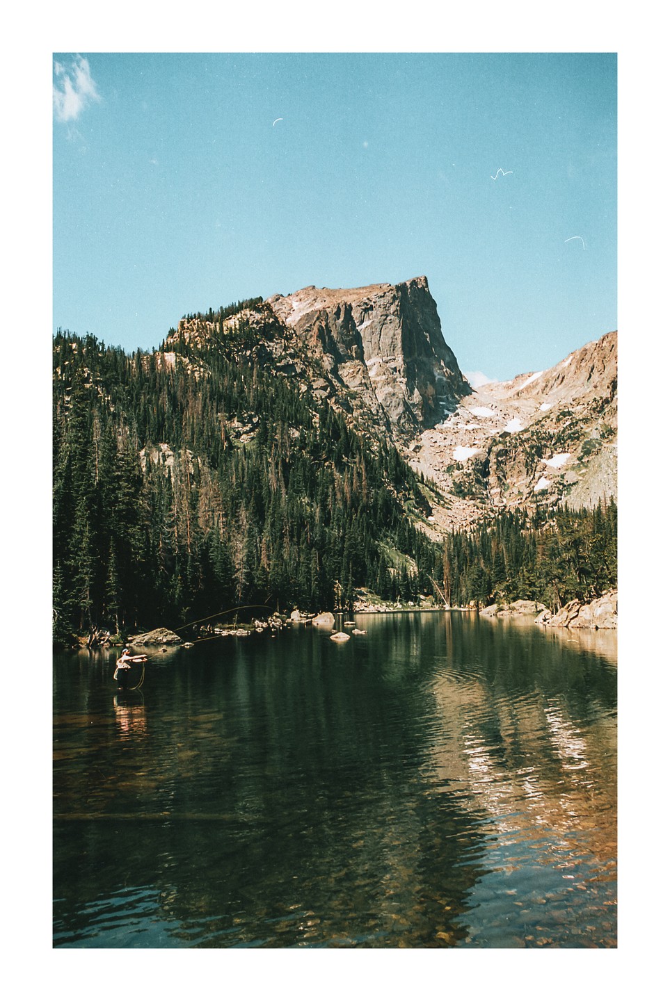 A man fishes in a mountain lake surrounded by pine trees and rocky peaks.
