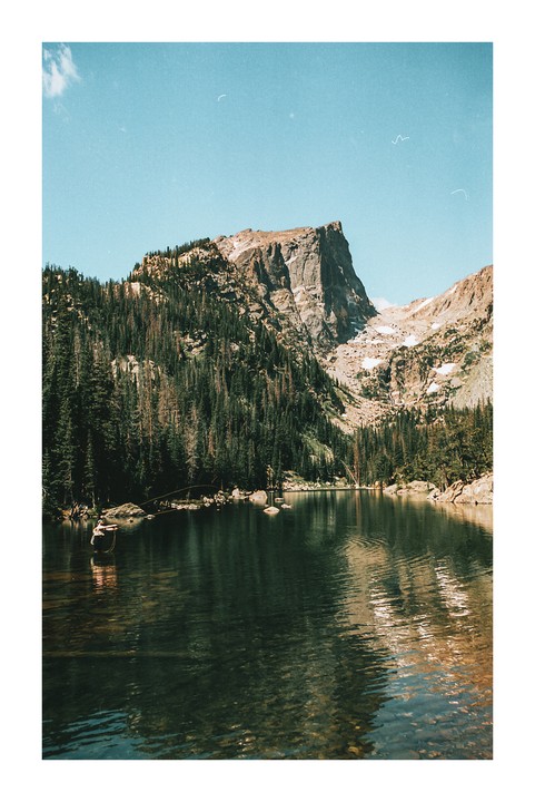A man fishes in a mountain lake surrounded by pine trees and rocky peaks.