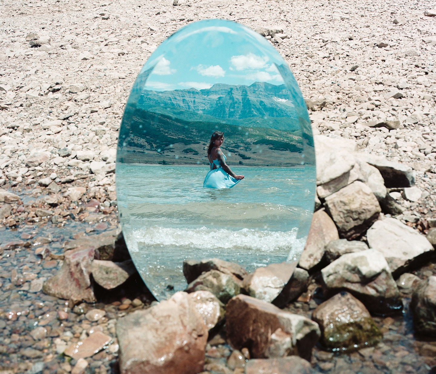 A woman in a blue dress stands in a lake, reflected in an oval mirror on a rocky shore.