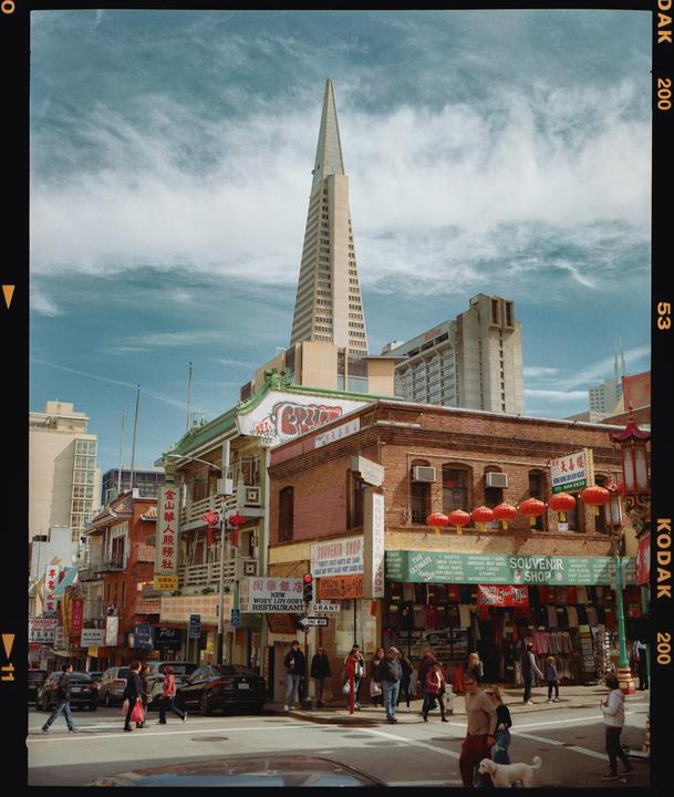 A street scene in San Francisco shows people walking on a sidewalk and crossing a street with buildings and a tall, pointed skyscraper in the background.