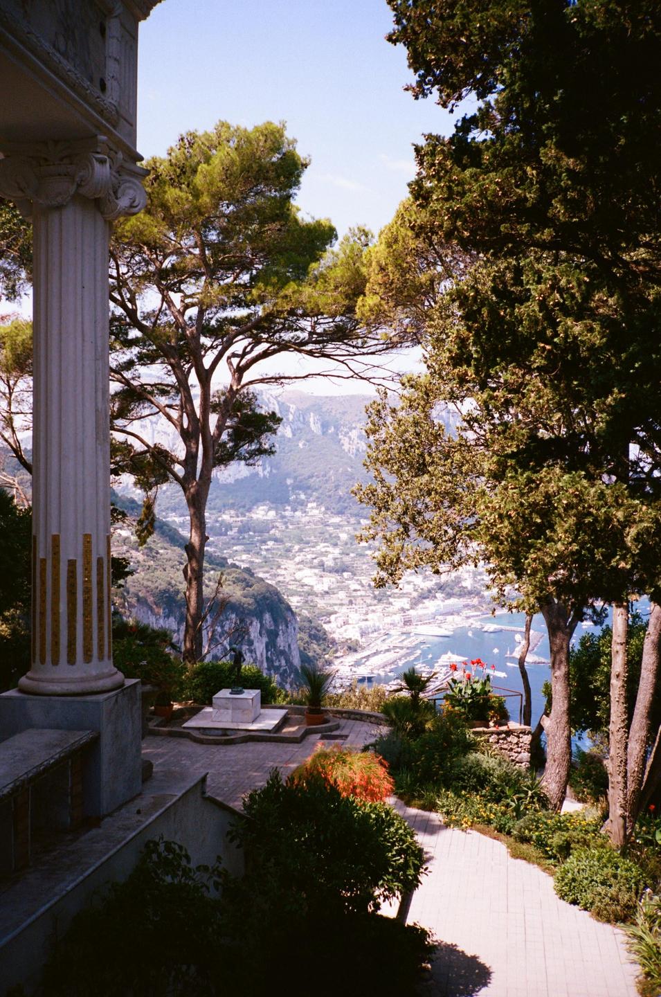 A scenic overlook features a classical column and lush trees framing a view of a coastal town and harbor below.