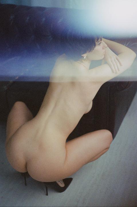 A nude woman crouches on the floor with her back to the viewer, looking to the right.
