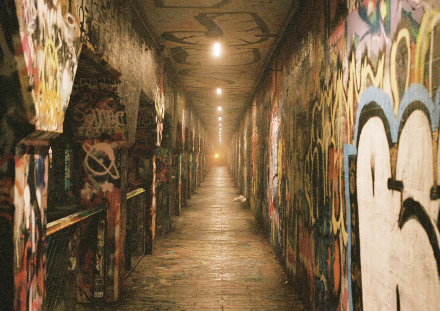 A long, narrow, graffiti-covered tunnel is illuminated by a series of lights on the ceiling, leading to a bright light at the far end.