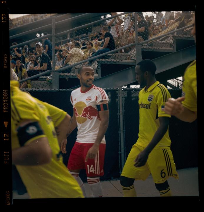 Three men in soccer uniforms are on a field with spectators in the background.