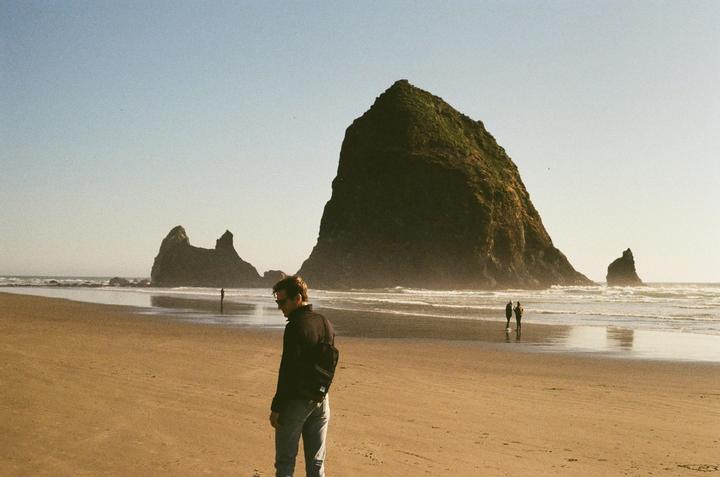 A man stands on a sandy beach with large rock formations in the background and the ocean to his right.