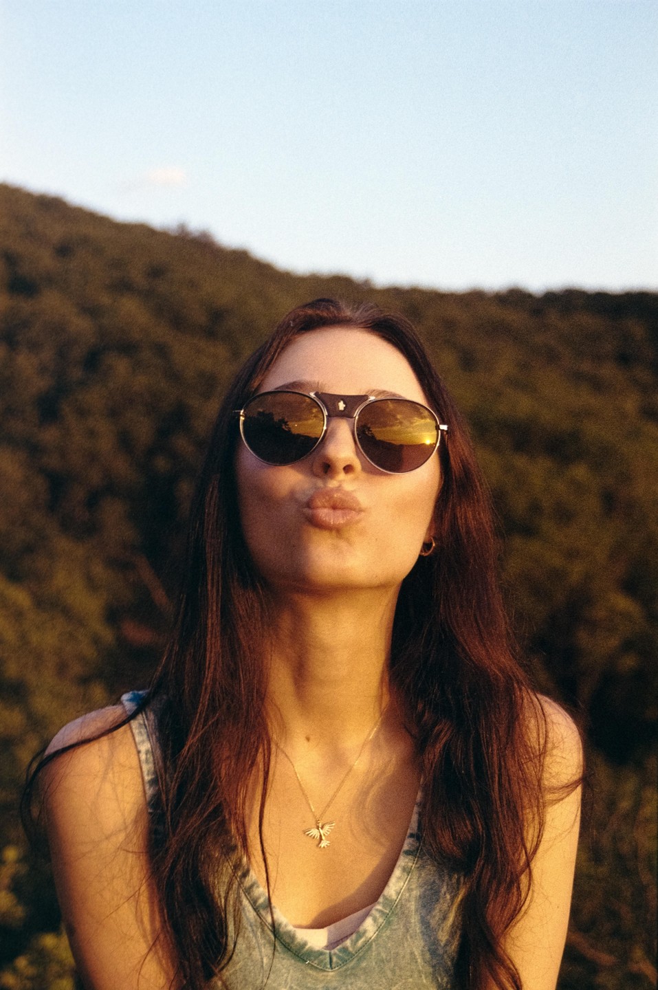 A woman wearing sunglasses and a denim top puckers her lips for a kiss, with a green hillside and clear sky in the background.