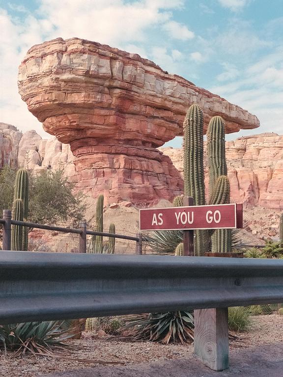 A red rock formation with a flat top stands behind a guardrail and a sign that reads "AS YOU GO" amidst cacti.
