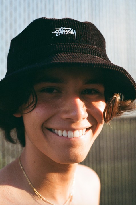 A young man wearing a bucket hat smiles at the camera with sunlight on his face.