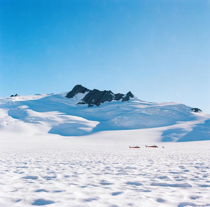 Two red helicopters are parked on a vast snowfield with a large snow-covered mountain in the background under a clear blue sky.
