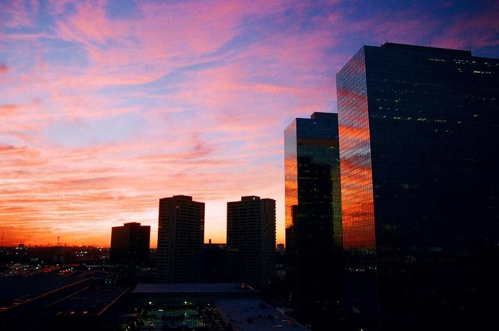 A vibrant sunset sky with pink and orange clouds is reflected in the glass facades of several tall buildings.