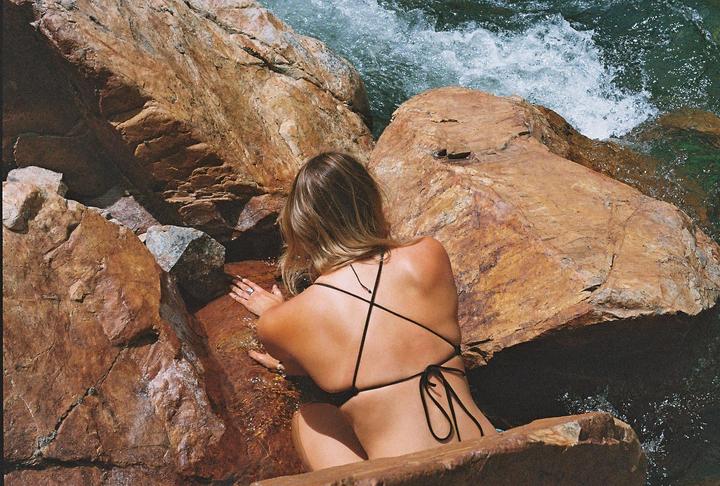A woman in a black bikini top is leaning on a large rock next to rushing water.