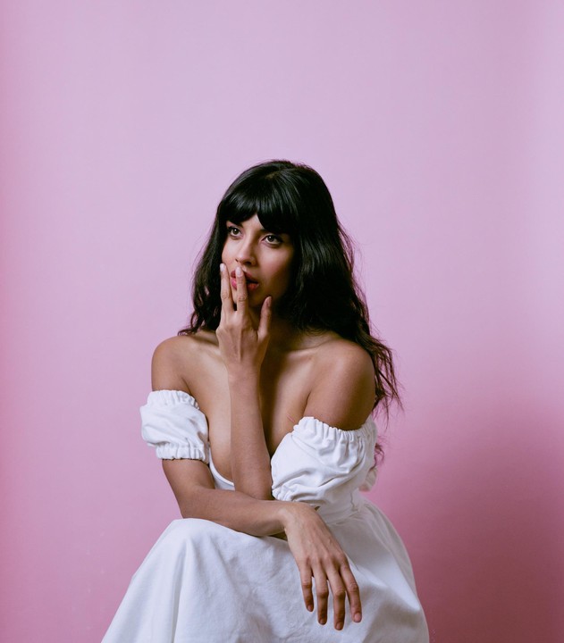 A woman in a white dress with dark hair looks up with her hand to her mouth against a pink background.