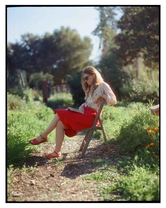 A woman wearing sunglasses and a red skirt sits on a wooden chair in a garden, reading a book.