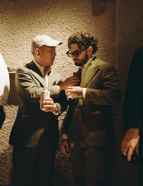 Two men in suits are talking to each other while one holds a can and the other holds a glass.