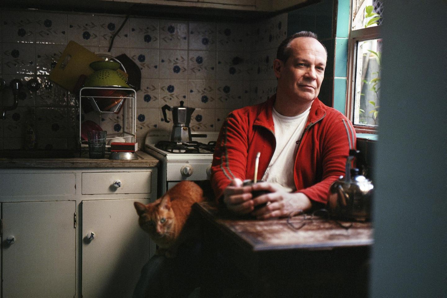 A man in a red jacket sits at a wooden table in a kitchen with a cat beside him.
