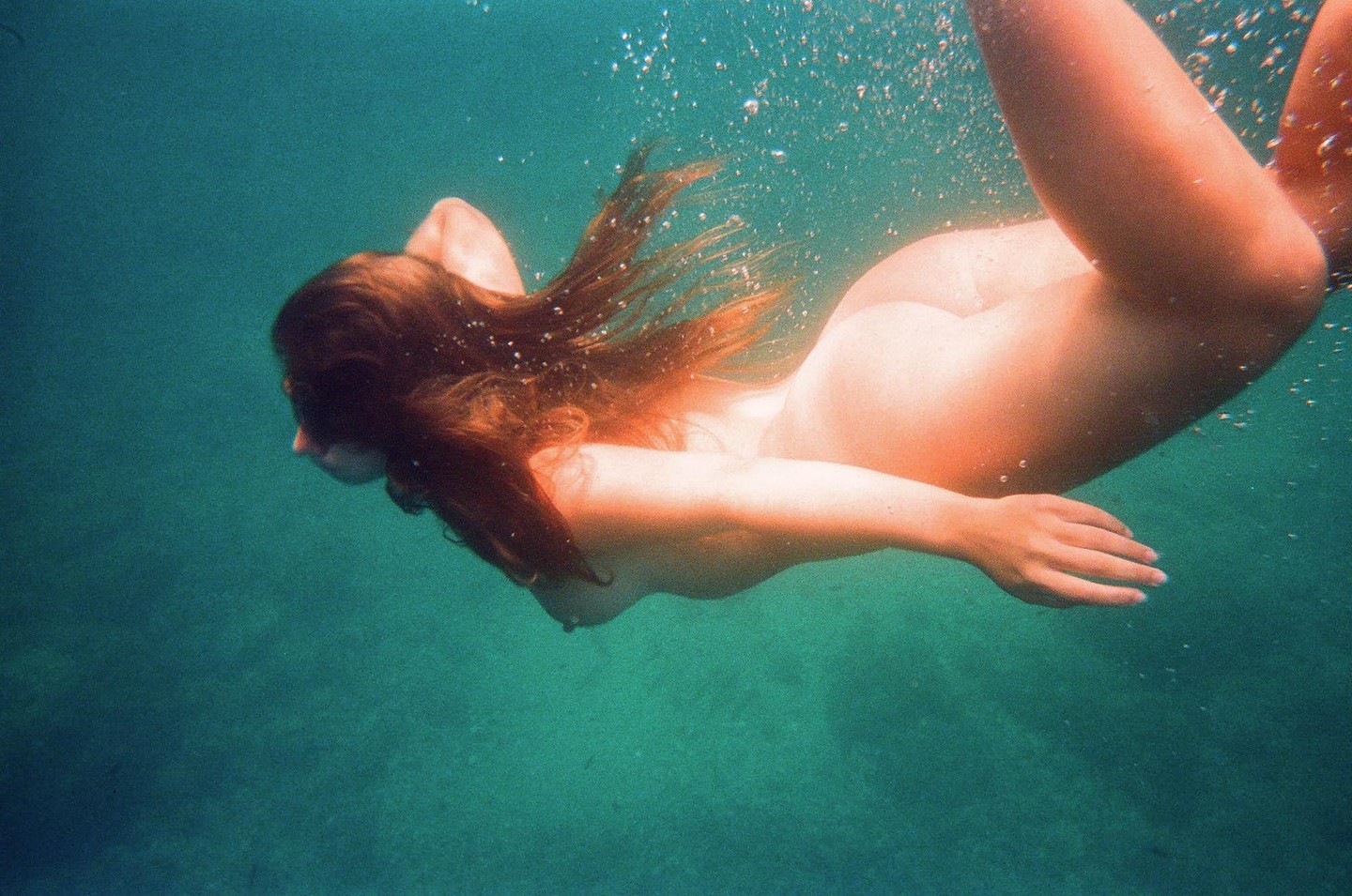 A woman with long hair swims nude underwater.