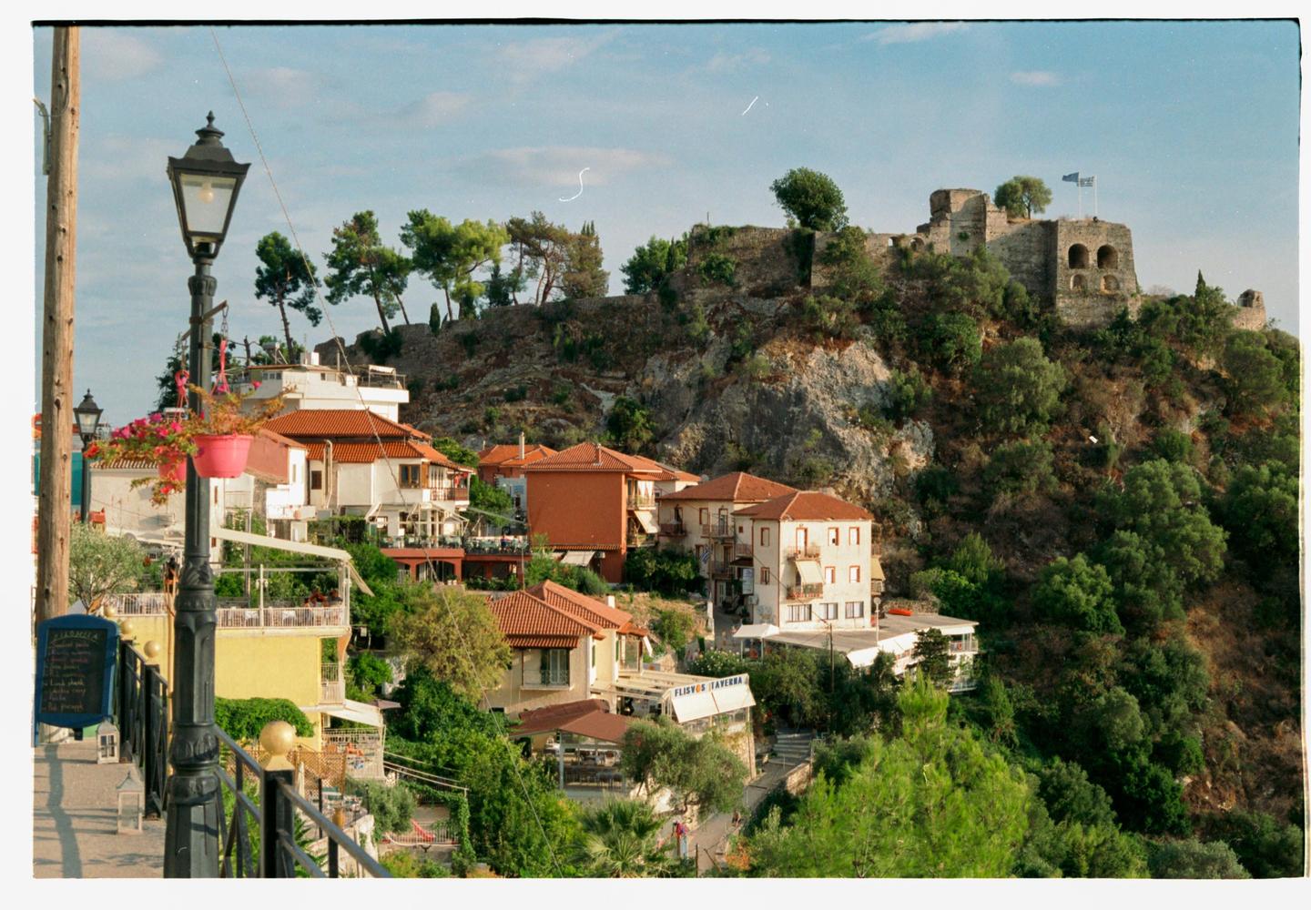 A village with red-roofed buildings is nestled on a hillside below a stone fortress.