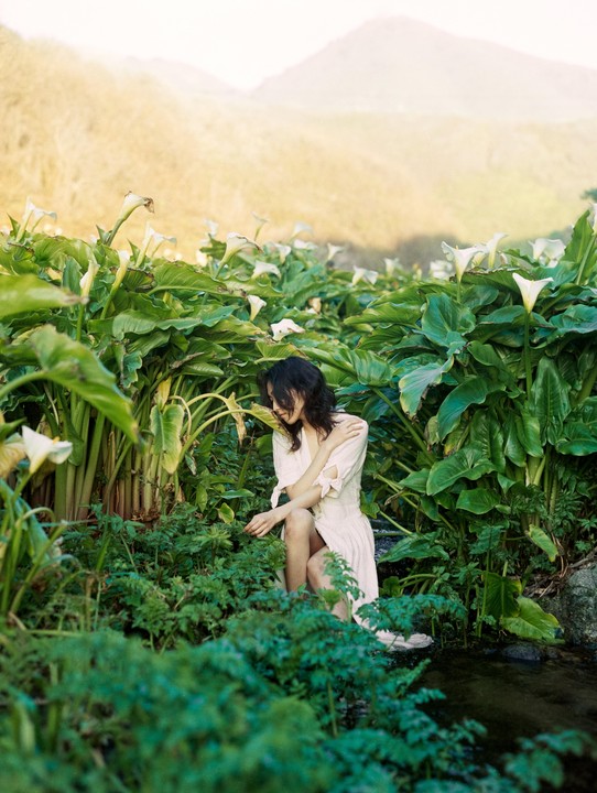 A woman in a white dress sits among calla lilies and other plants next to a stream with mountains in the background.