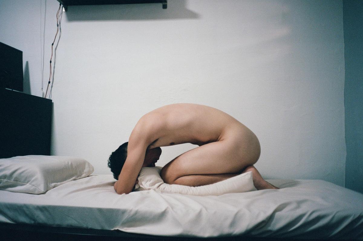 A nude man kneels on a bed with his head resting on a pillow.