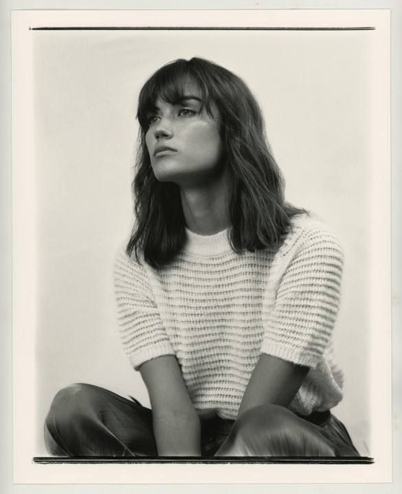 A woman with bangs and long hair sits looking to the left in a studio portrait.