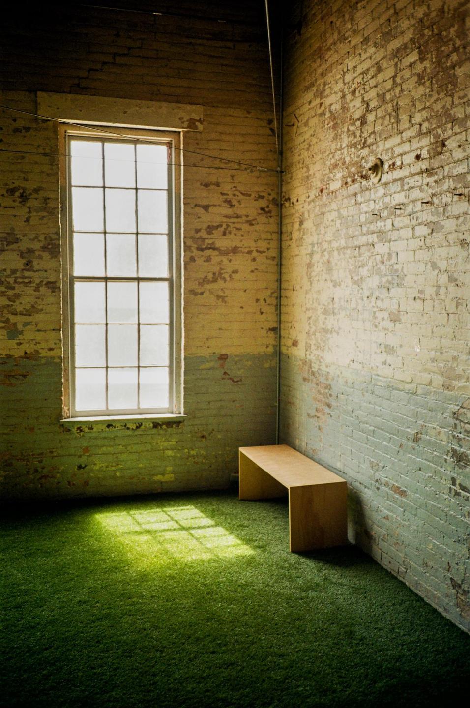 Sunlight streams through a tall window onto a green carpeted floor next to a wooden bench in a room with distressed brick walls.