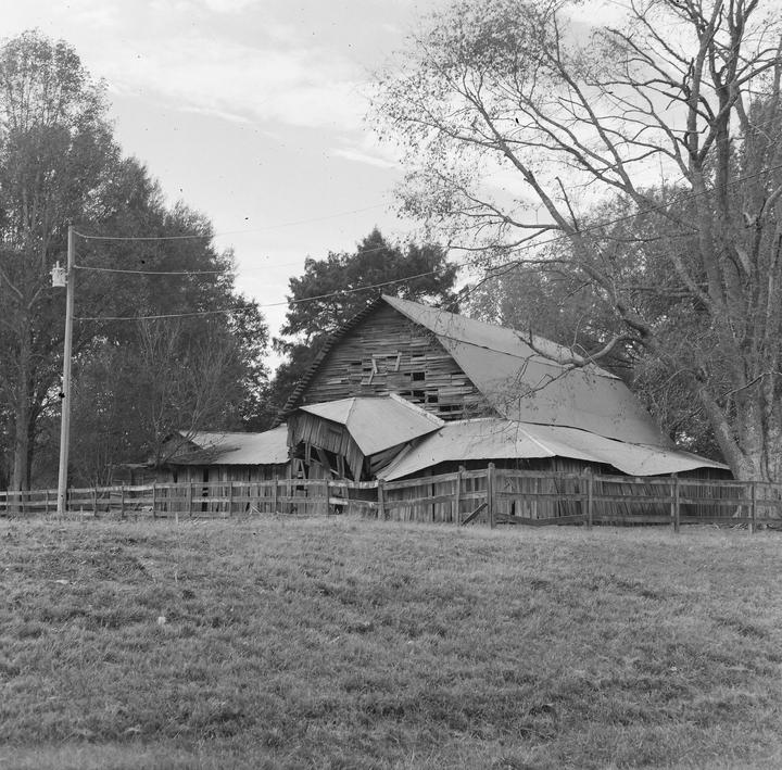 An old wooden barn with a collapsing roof stands behind a wooden fence and a grassy field.