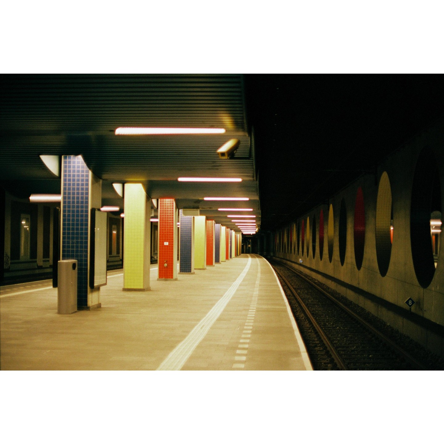 A subway platform with colorful pillars and a train track is illuminated by overhead lights.