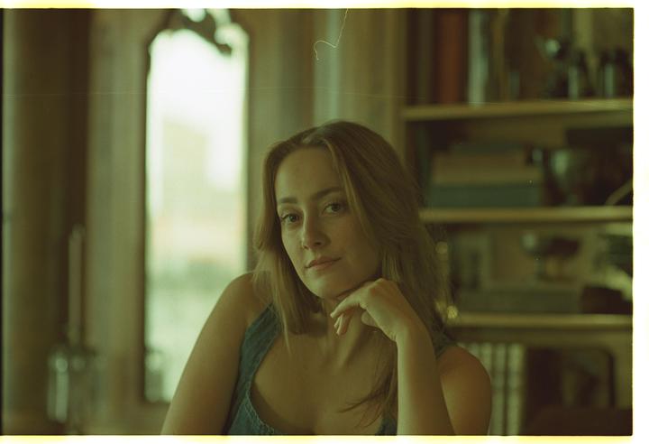 A woman with long hair and a nose ring rests her chin on her hand, looking directly at the viewer.