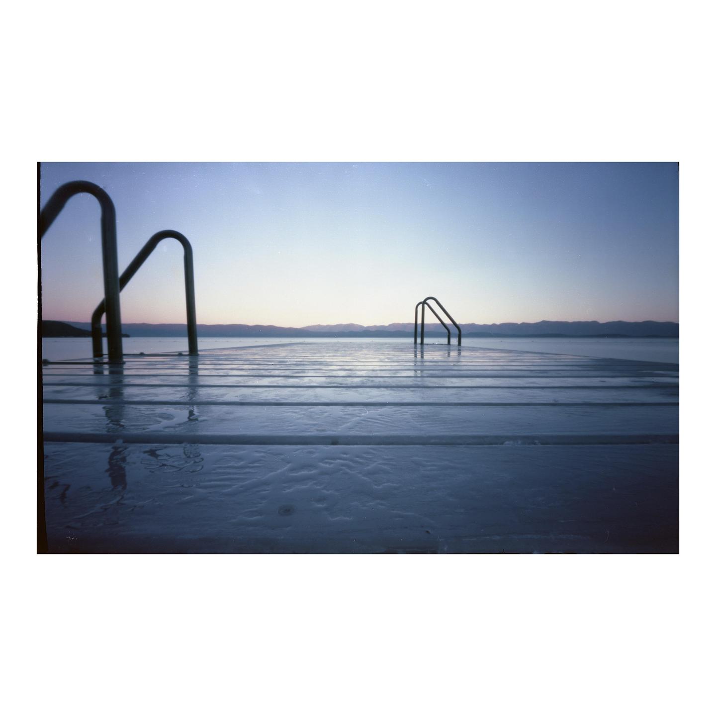 A wooden dock with two sets of metal steps leading into a body of water is shown under a clear sky with distant mountains.