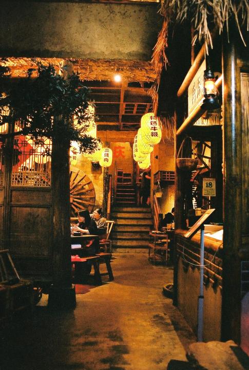 A restaurant interior is illuminated by hanging lanterns and warm light, with people seated at tables and a staircase leading upwards.