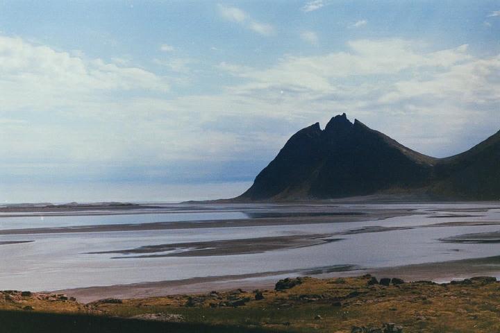 A vast landscape features a large mountain range behind a body of water with a grassy foreground under a cloudy sky.