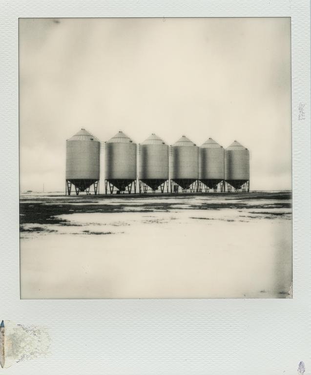 Six grain silos stand in a row on a flat, snow-covered landscape under a bright sky.
