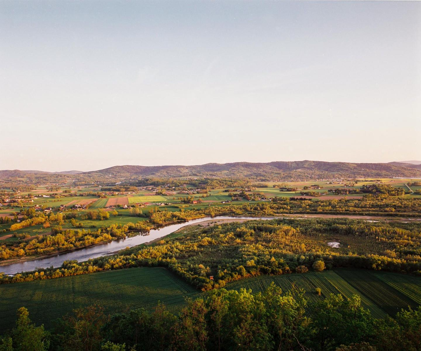 A river winds through a green valley with fields and houses under a clear sky.