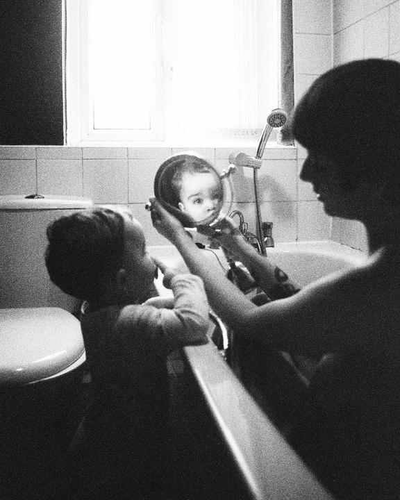 A child in a bathtub holds a mirror reflecting a baby's face.