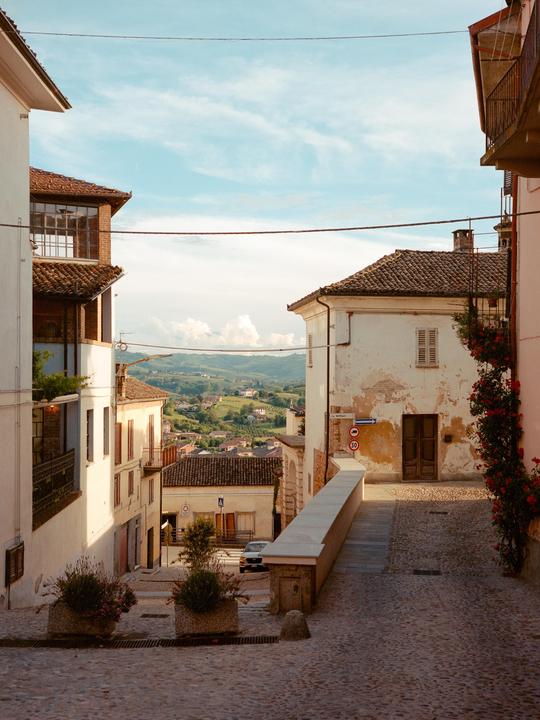 A narrow cobblestone street in a village is lined with buildings and leads to a view of rolling hills and distant houses.