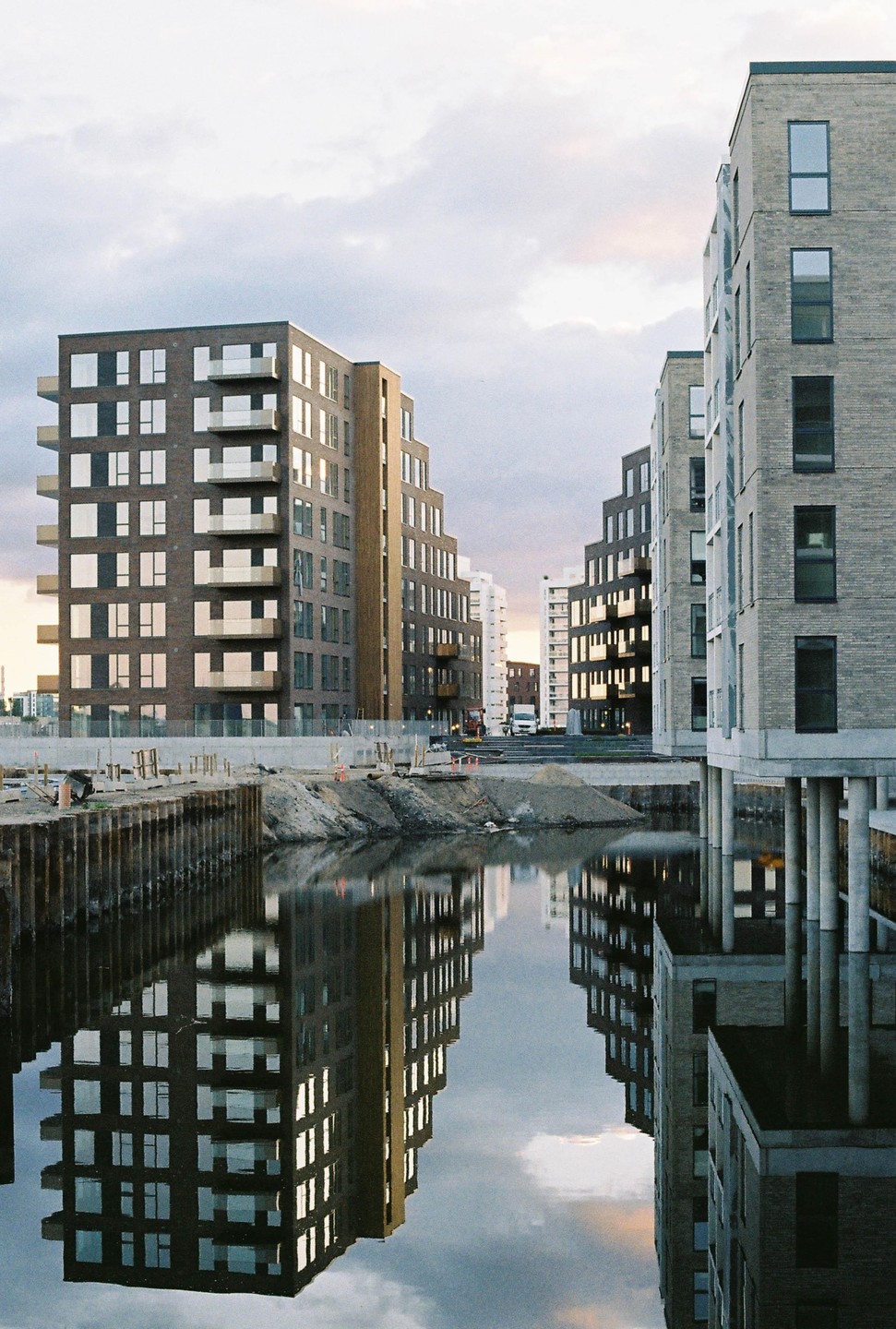 Modern buildings with balconies are reflected in the water of a canal next to a construction site.