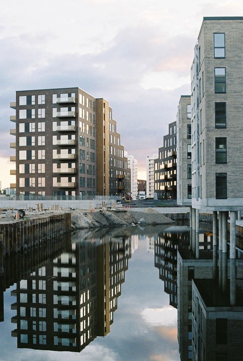 Modern buildings with balconies are reflected in the water of a canal next to a construction site.