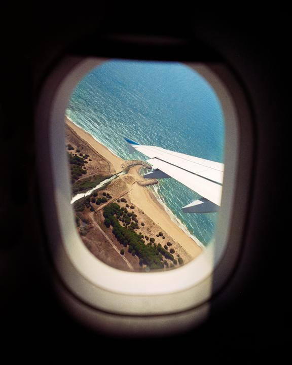 An airplane wing is visible through a window, flying over a coastline with a beach and ocean.