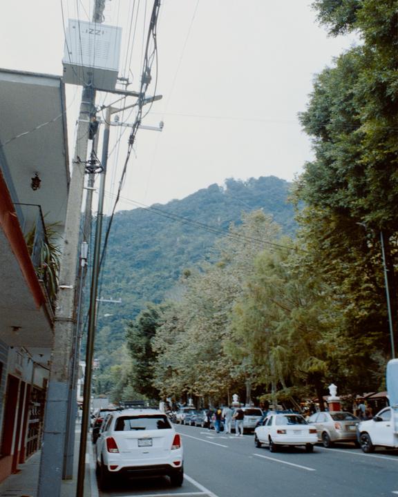 A street in Orizaba, Mexico, is lined with parked cars and trees, with a mountain in the background.
