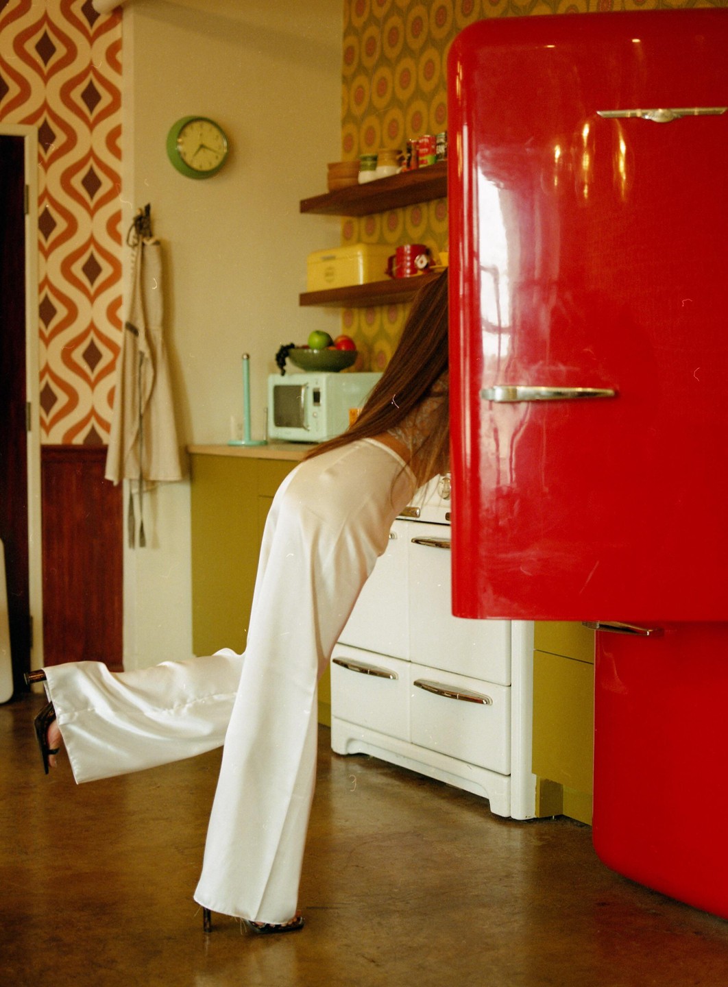 A woman in white pants bends over to look inside a red refrigerator in a kitchen with patterned wallpaper.