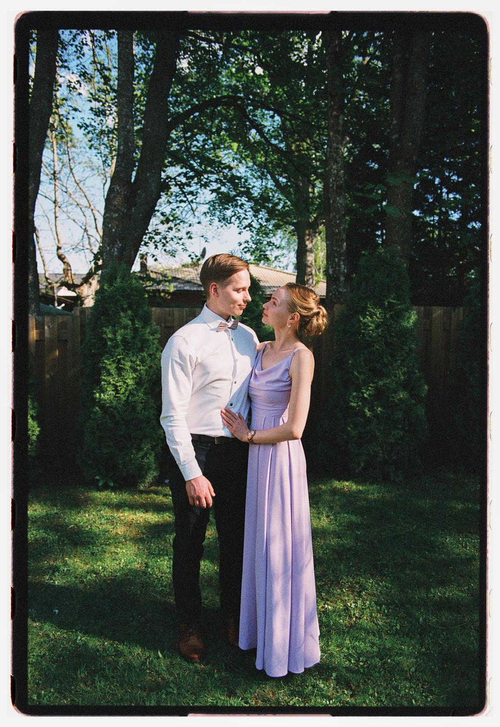 A couple stands in a grassy yard surrounded by trees and bushes.