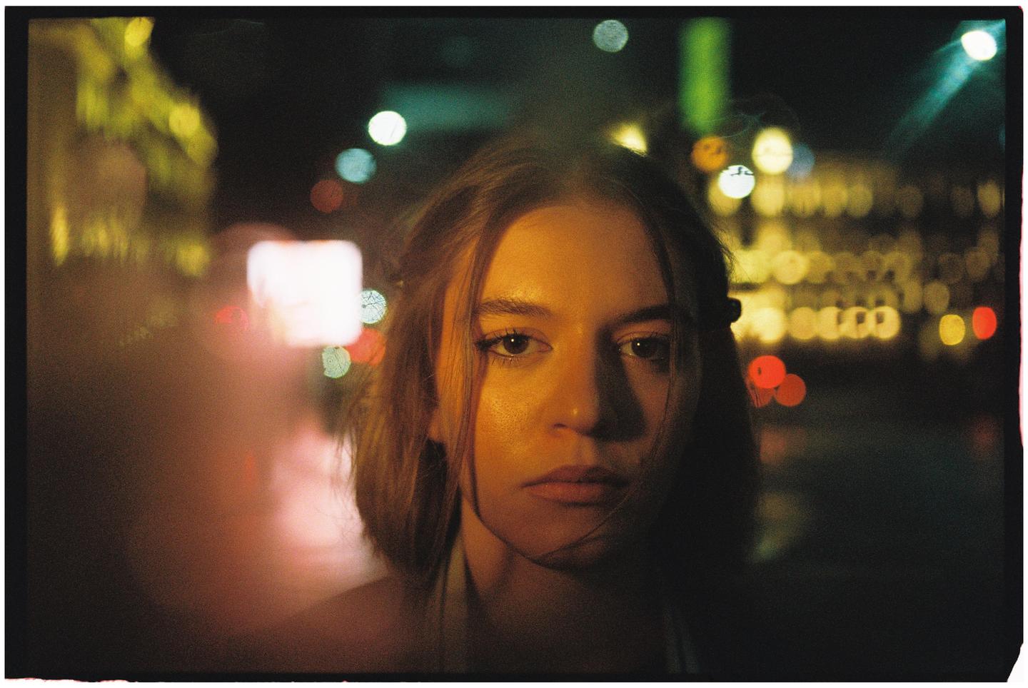 A woman with dark hair looks directly at the viewer with blurred lights in the background.