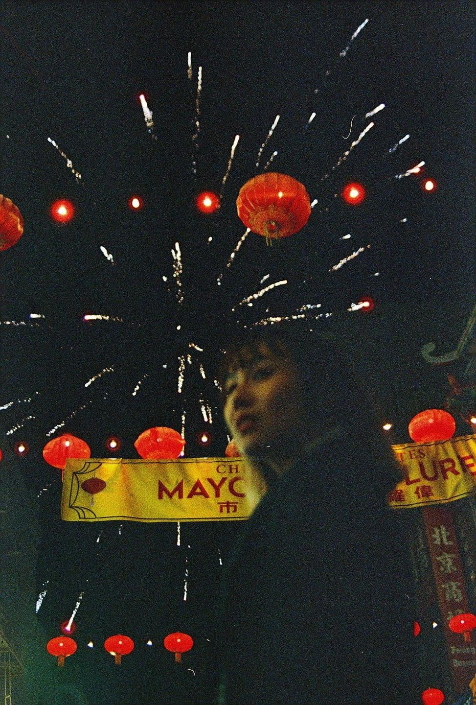 A woman is visible in front of fireworks and red lanterns at night.